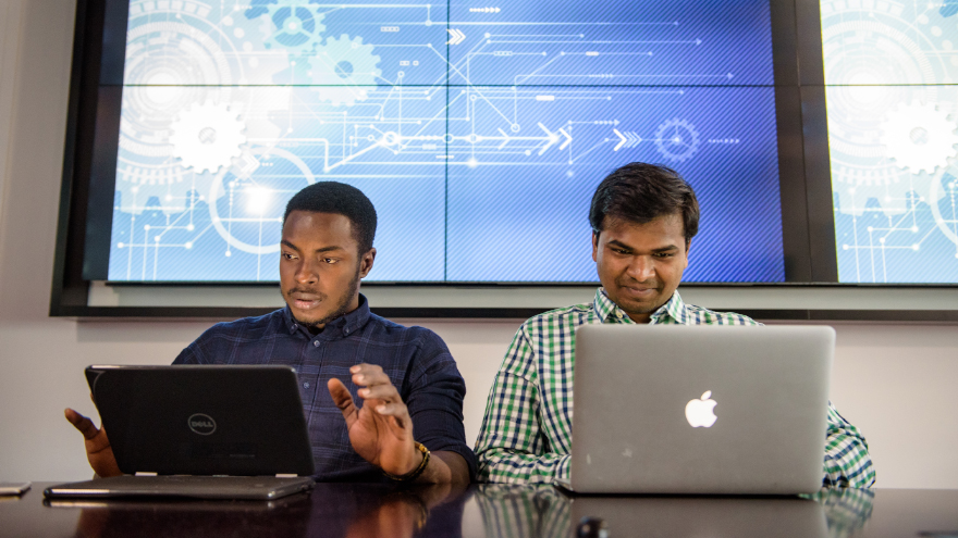 Two students sit in front of laptops with an engineering graphic on a screen behind them.