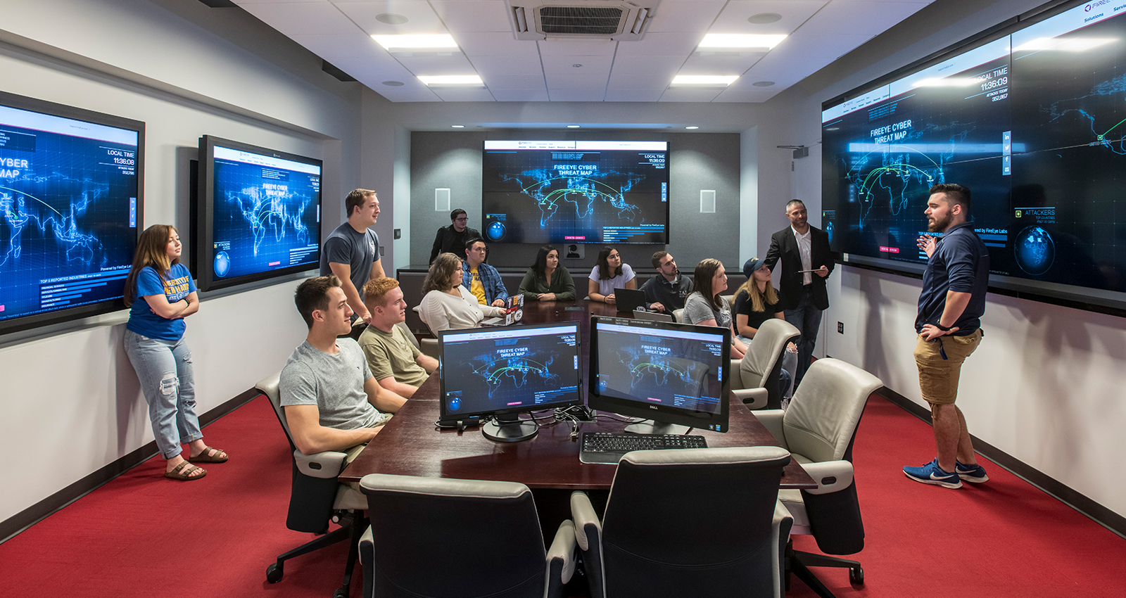 A presenter stands in front of a large display showing a global cyber threat map, speaking to a group of students seated around a conference table in a high-tech cybersecurity operations center.