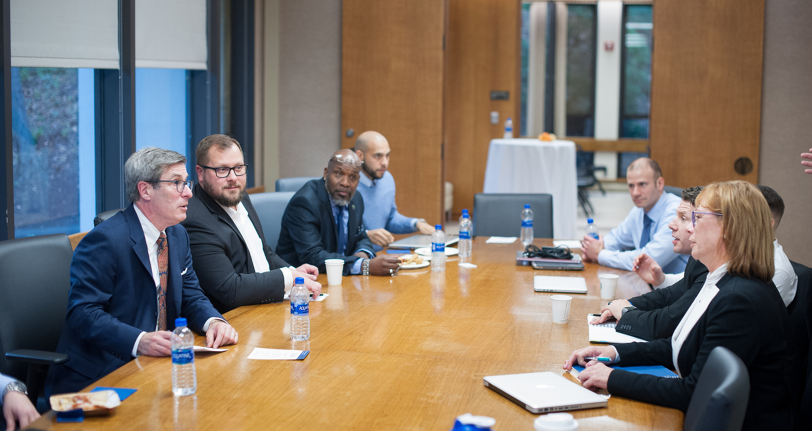 A group of professionals in business attire seated around a wooden conference table during a meeting in a boardroom setting.