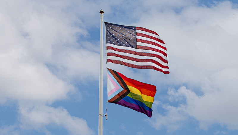 An American flag and an LGBTQ+ flag waving on a flag pole.