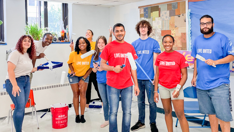 University of New Haven volunteers for the United Way painting a classroom at West Haven Child Development Center. Students holding paint brushes look at the camera and smile, each wearing a red or blue Live United shirt.