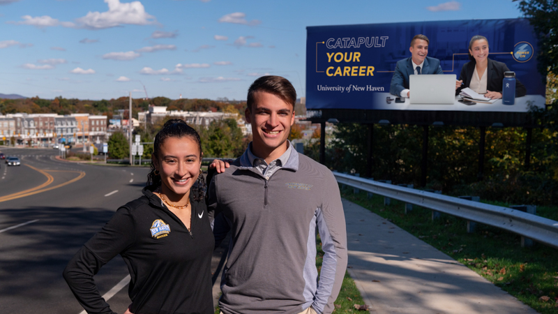 Two students stand by a Power On billboard where they are also featured